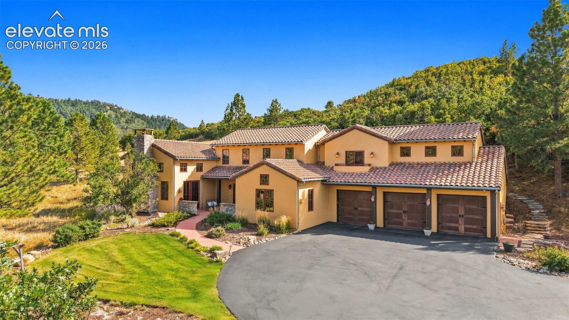 Stucco exterior with a tiled roof, three-car garage, expansive paved driveway, and established landscaping with mature trees