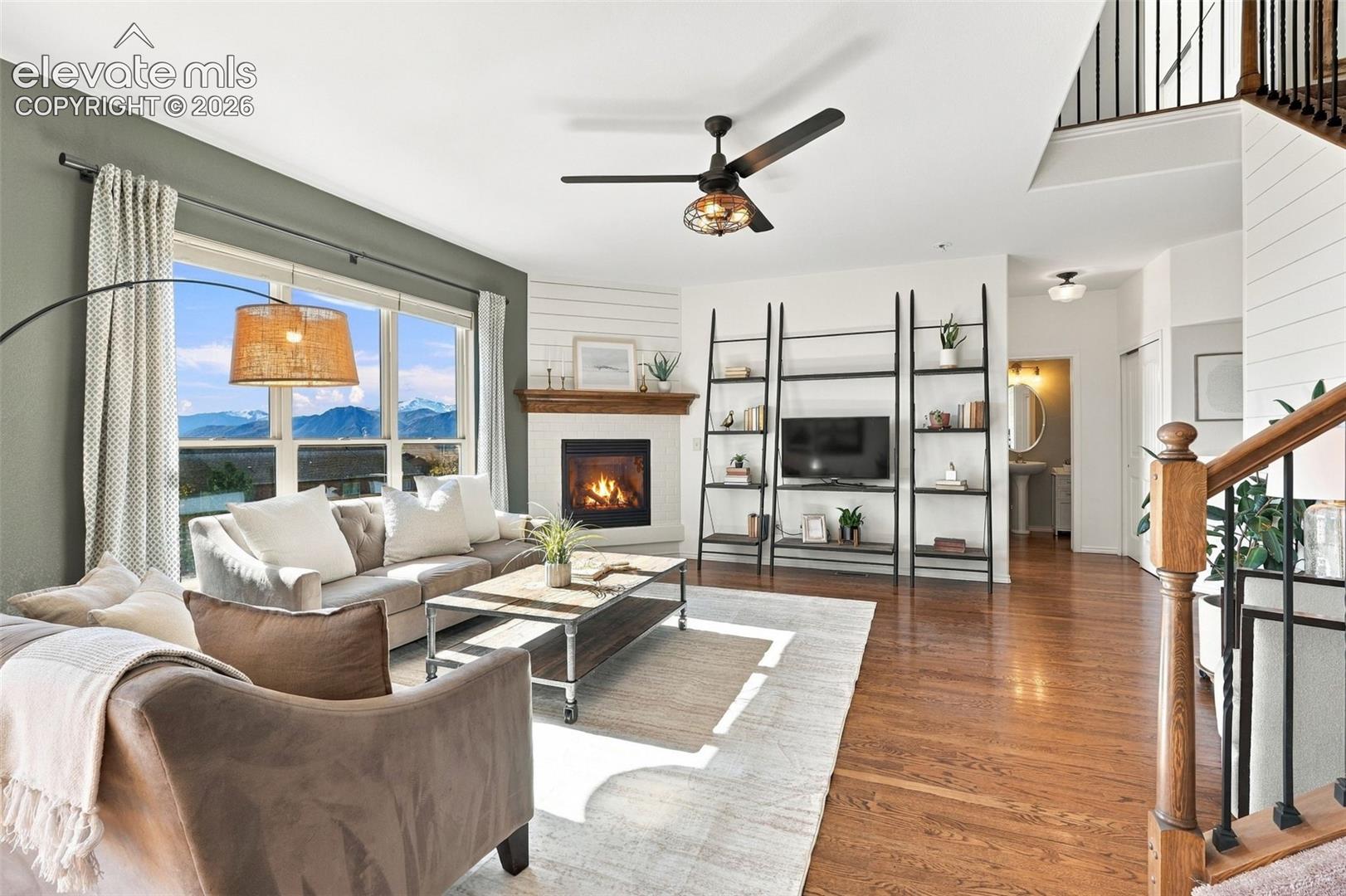 Open-concept living space featuring wood-finish flooring, corner fireplace with a wooden mantel, white shiplap wall accents, and an open staircase with a wooden handrail and metal balusters