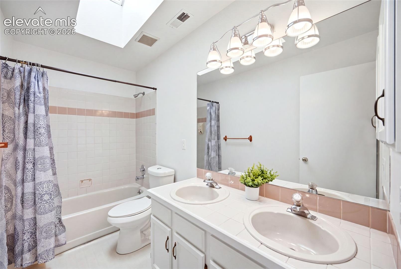 Bathroom featuring a double-sink vanity with tile countertops, a large vanity mirror with multi-bulb light fixture, a skylight, and a porcelain-tiled tub and shower combination