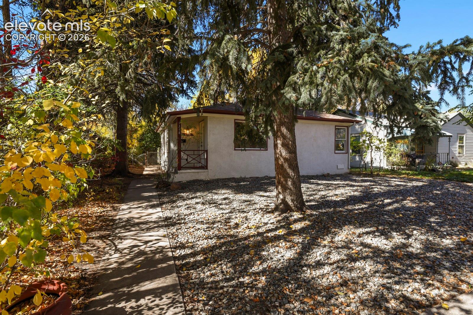 Stucco exterior residence featuring a covered front porch, concrete walkway, and low-maintenance rock landscaping with mature coniferous trees