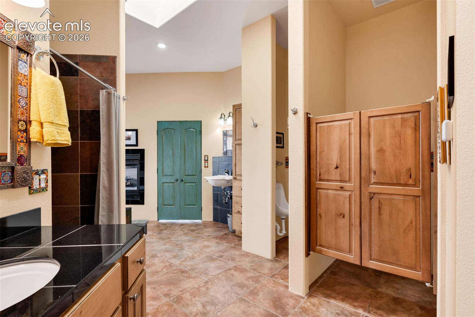 Bathroom featuring tile flooring, a dark stone vanity countertop with an integrated basin, a stall enclosed by wood cabinet-style doors, a wall-mounted urinal, and a skylight