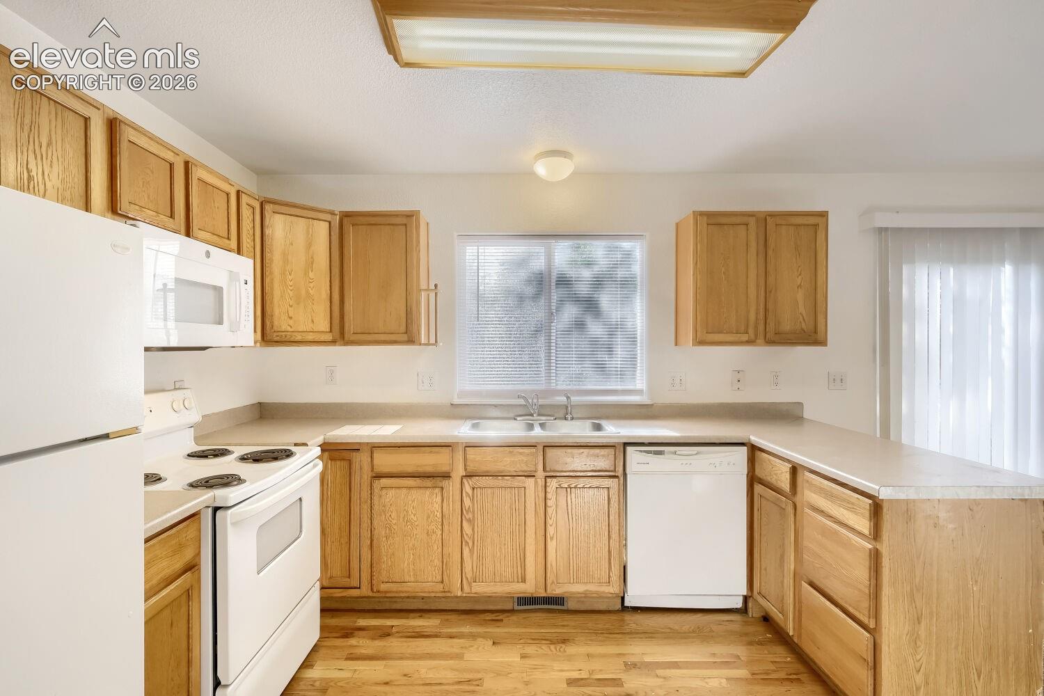 Kitchen featuring natural wood cabinetry, white appliances, dual-basin sink, and wood-finish flooring