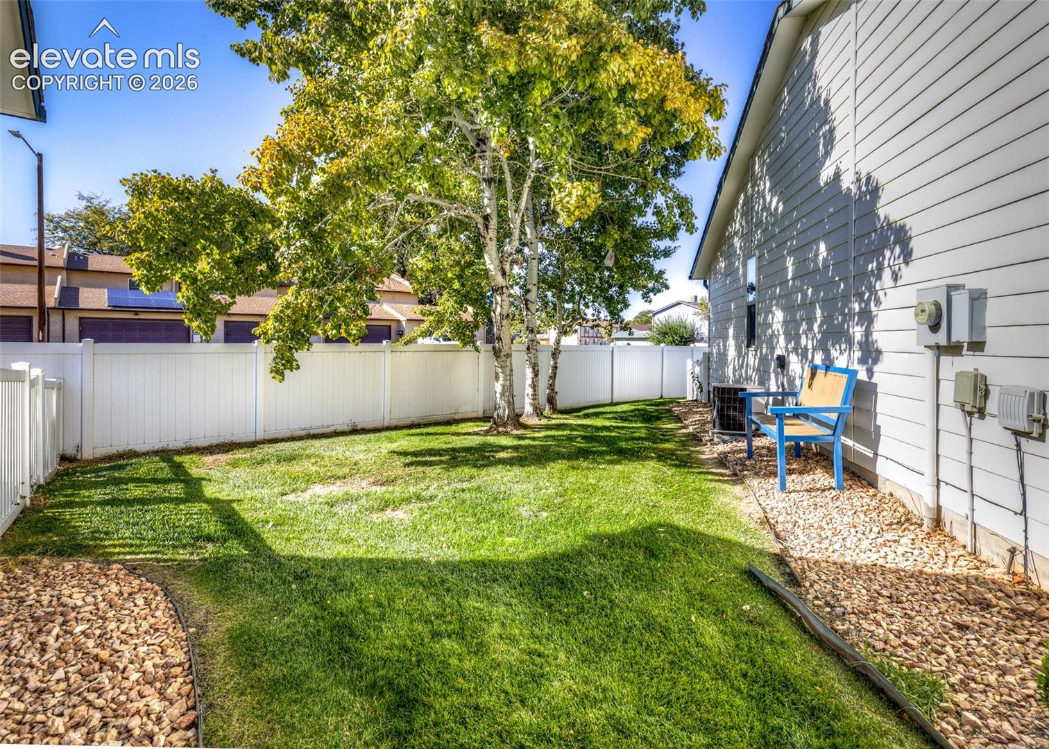 Side yard featuring horizontal lap siding, white vinyl fencing, a manicured lawn, mature trees, and a decorative rock ground cover