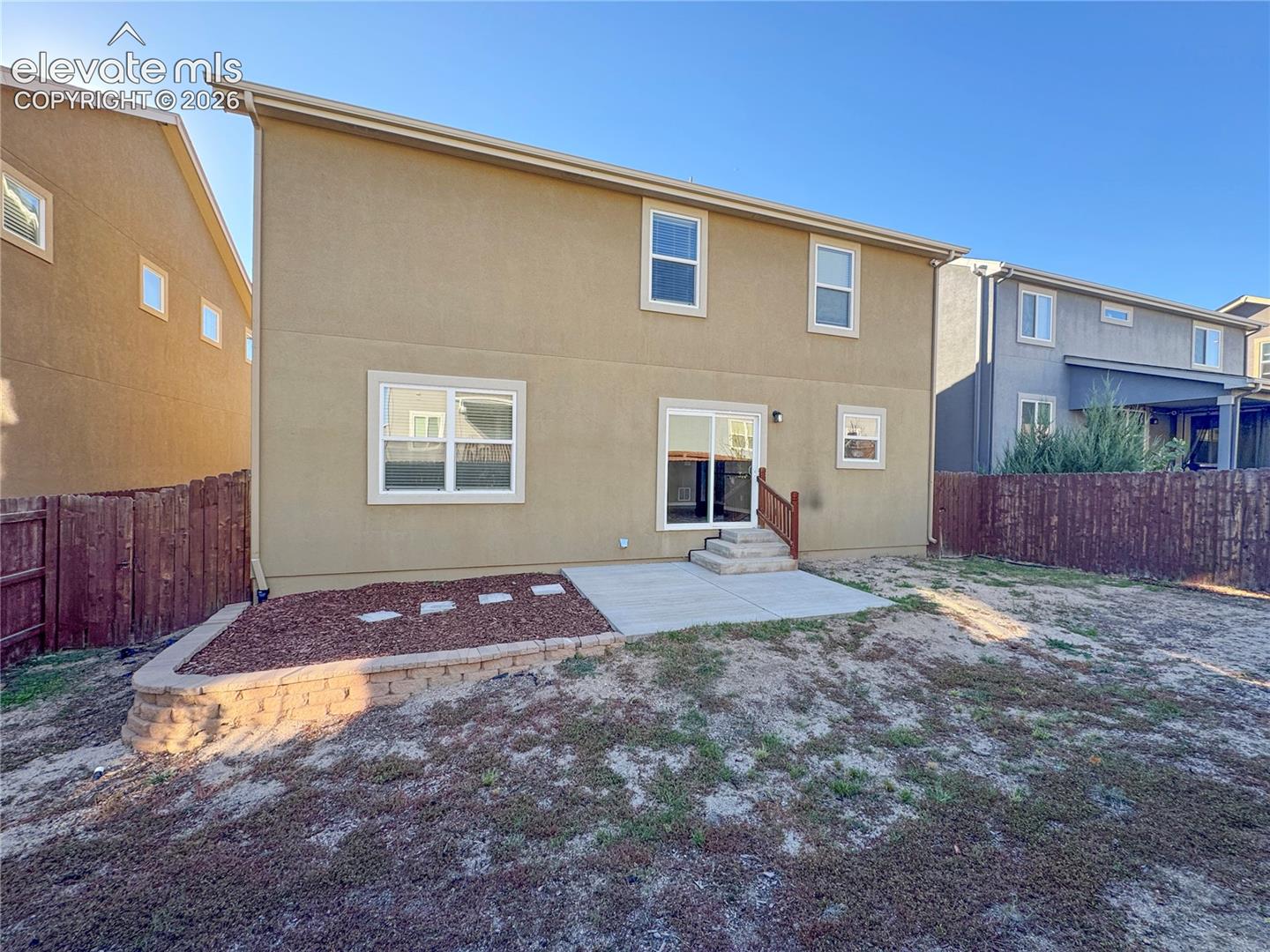 Stucco exterior featuring a concrete rear patio, sliding glass door, tiered masonry retaining wall, and wood-style privacy fencing