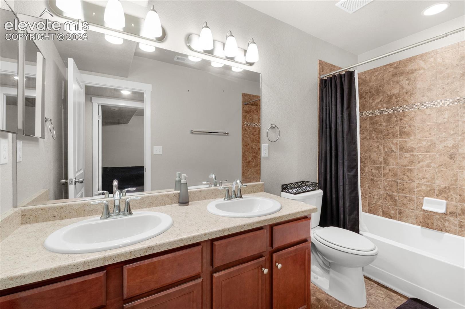 Dual-sink vanity with a solid-surface countertop, wood-finish cabinetry, chrome fixtures, and a tub-shower combination featuring decorative stone-tile surround