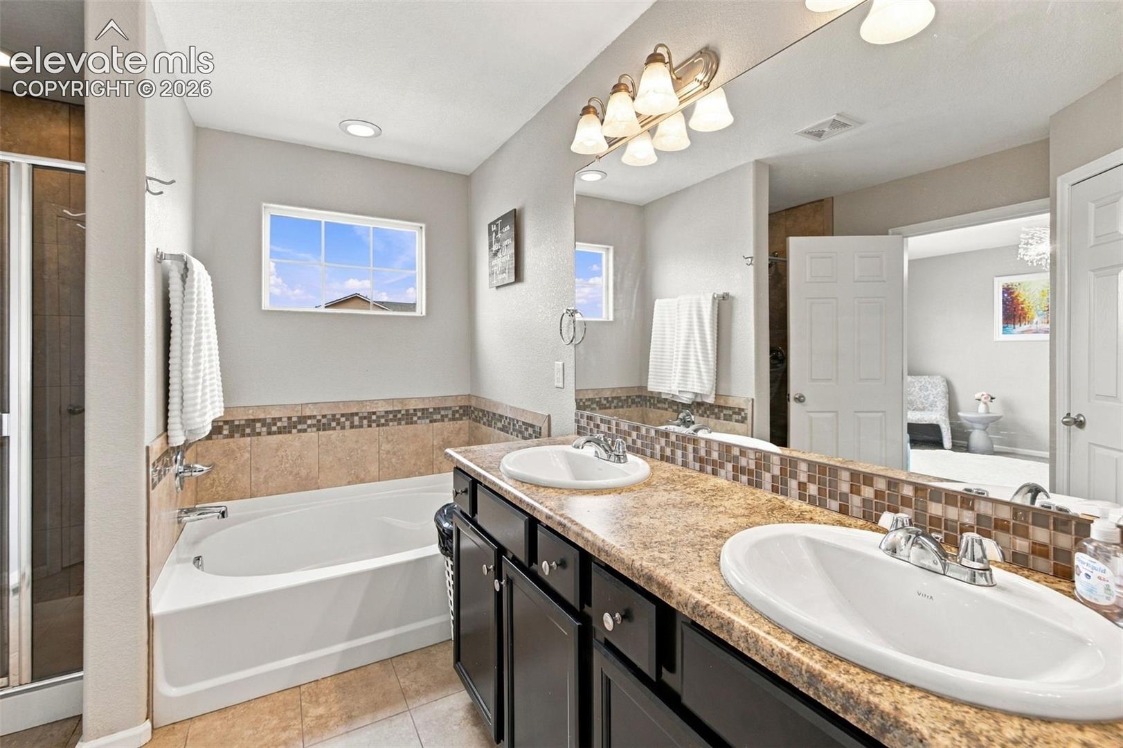 Dual-sink vanity with dark cabinetry, stone-finish countertop, soaking tub, mosaic tile backsplash, and full-width vanity mirror