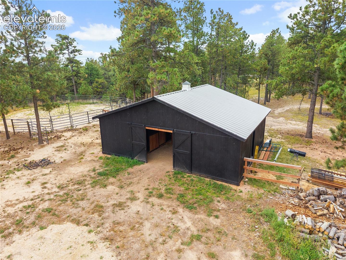Detached outbuilding featuring a metal roof, exterior sliding barn doors, and surrounding metal perimeter fencing on a wooded lot