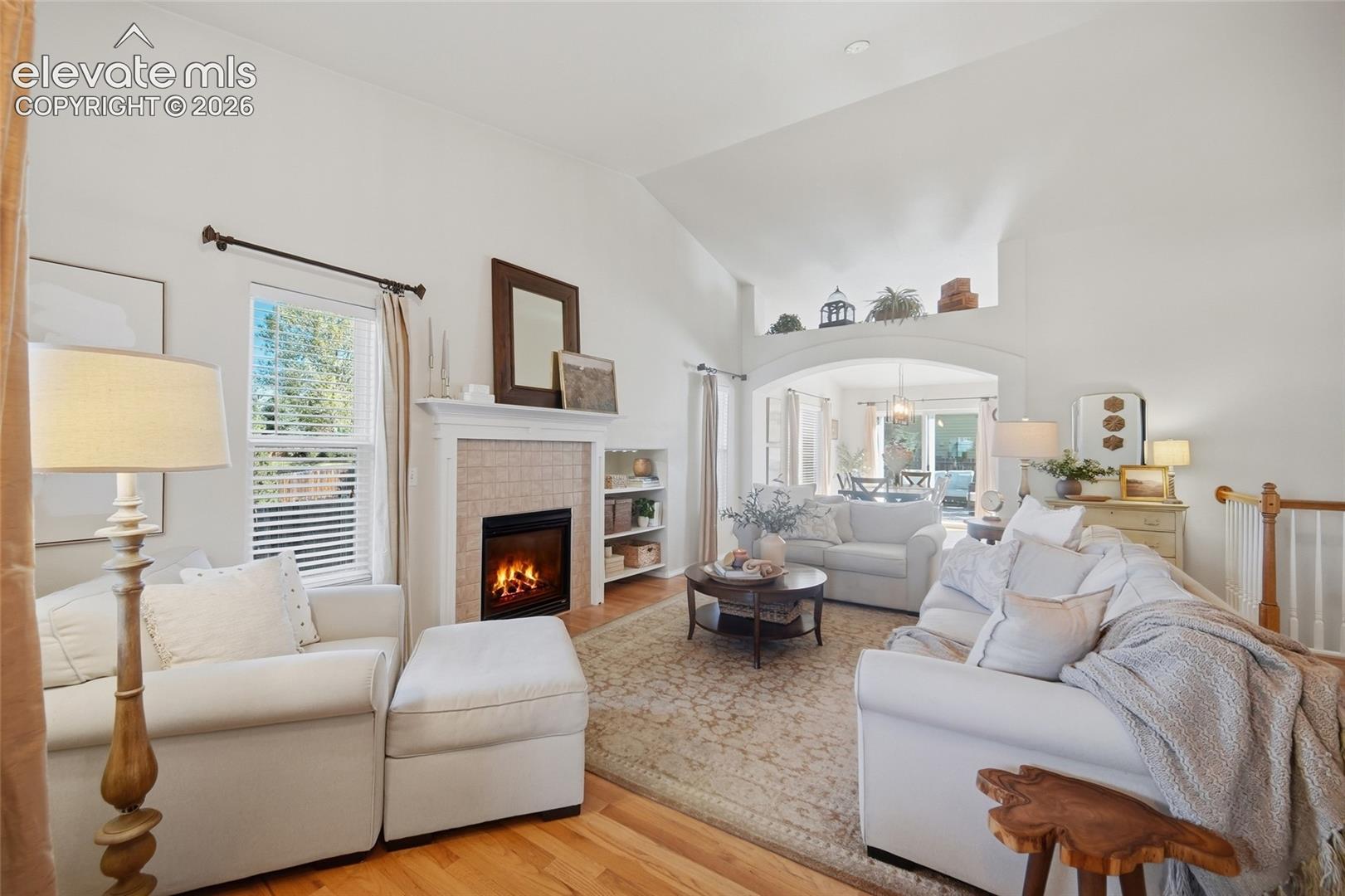 Vaulted ceilings with an arched architectural opening, wood-finish flooring, a fireplace with a tiled surround, and built-in shelving