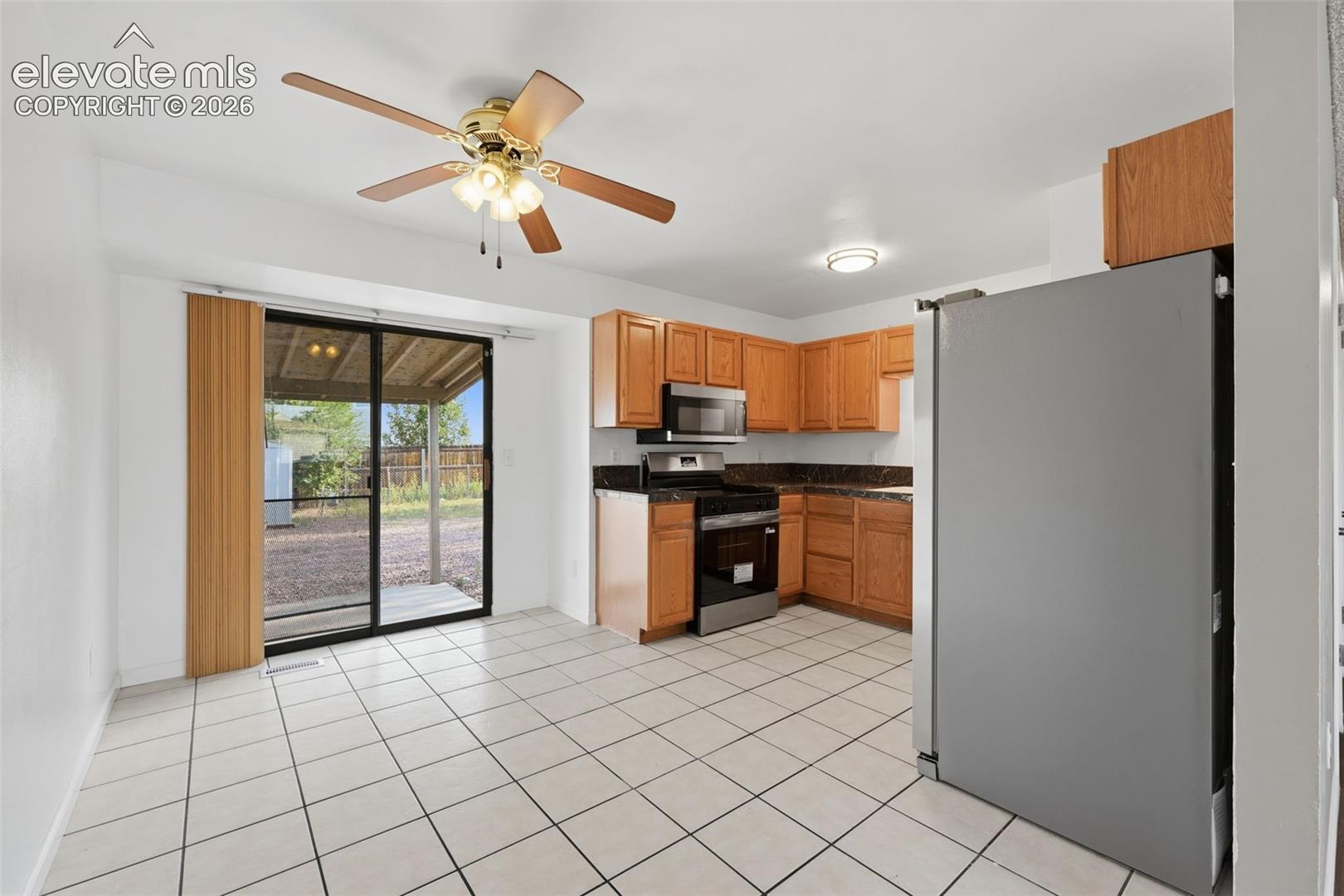 Kitchen featuring wood-finish cabinetry, dark countertops, stainless steel appliances, and white tile flooring