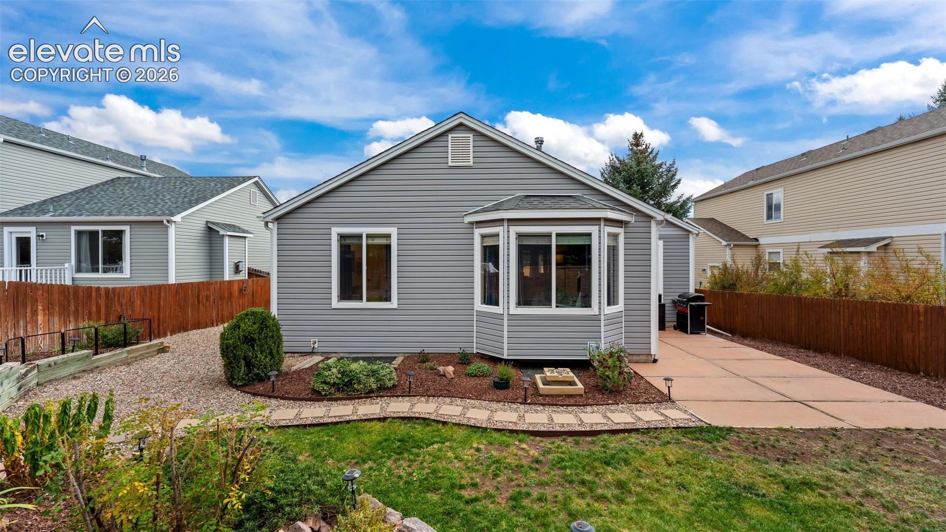 Exterior view featuring gray horizontal siding, a rear bay window, a paved concrete patio, a stone-path garden border, and wood privacy fencing