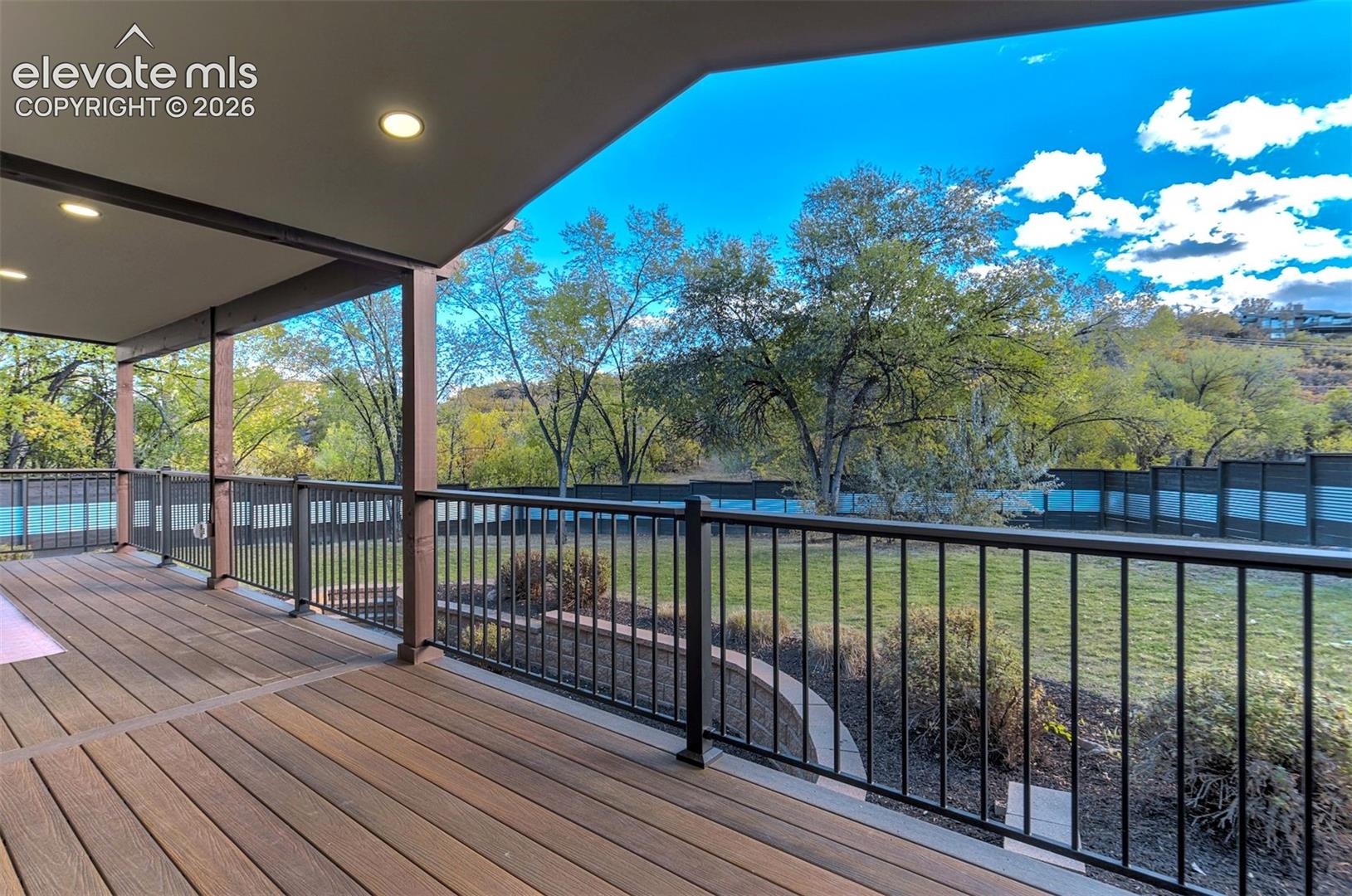Covered outdoor deck featuring wood-finish composite decking, recessed lighting, and black metal railings overlooking a backyard with mature trees and a tiered landscape wall