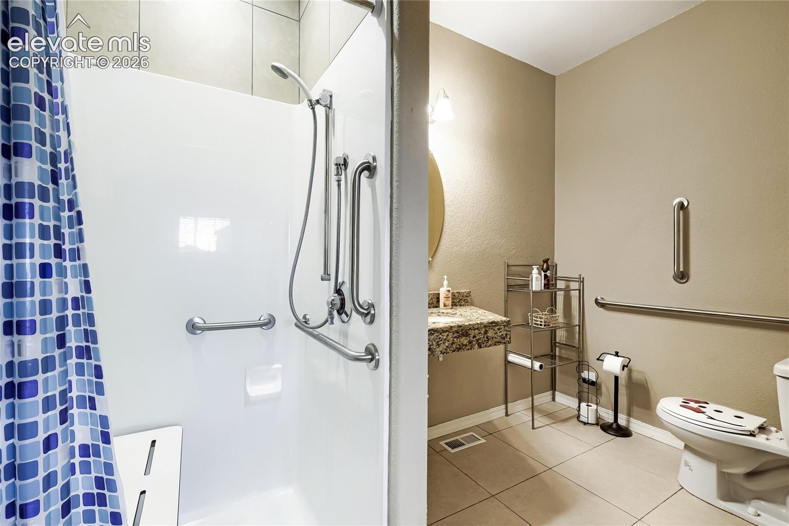This bath adjoins the main level bedroom-shower with grab bars and built-in seating, complemented by a vanity with a stone-finish countertop and an oval mirror