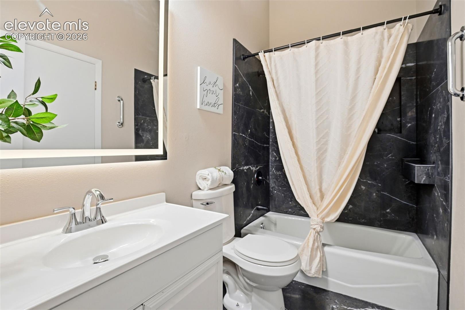 Bathroom with a vanity featuring an integrated sink and chrome faucet, an illuminated mirror, and a tub with dark marble-patterned surround