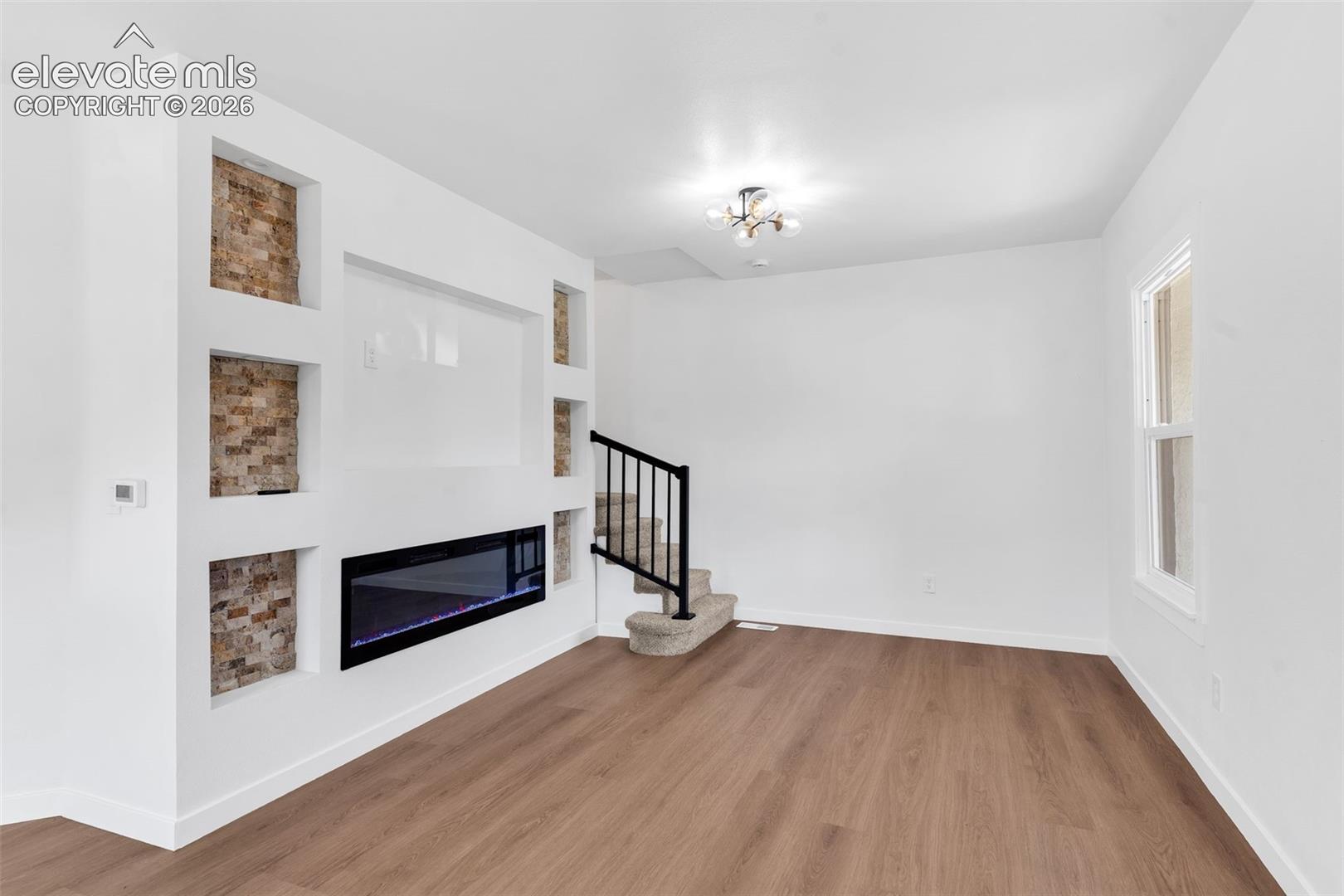 Living area featuring a linear electric fireplace, stone-accented built-in shelving, wood-finish flooring, a multi-arm flush-mount light fixture, and a staircase with a black metal railing