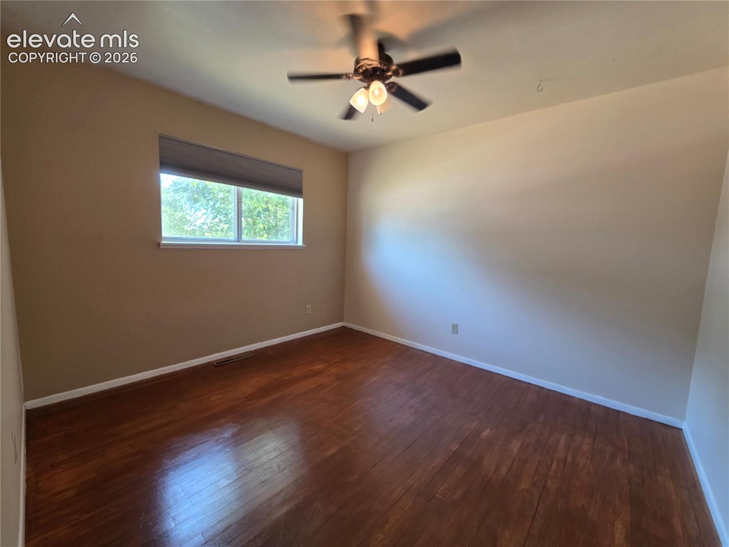 Room featuring wood-finish flooring, a ceiling fan with integrated lighting, and a window with roller shade