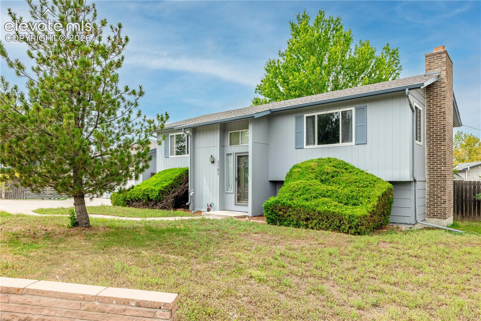 Split-level exterior featuring light-gray horizontal siding, a prominent brick chimney, multi-paned windows, and a landscaped front yard with mature pine trees and shrubs