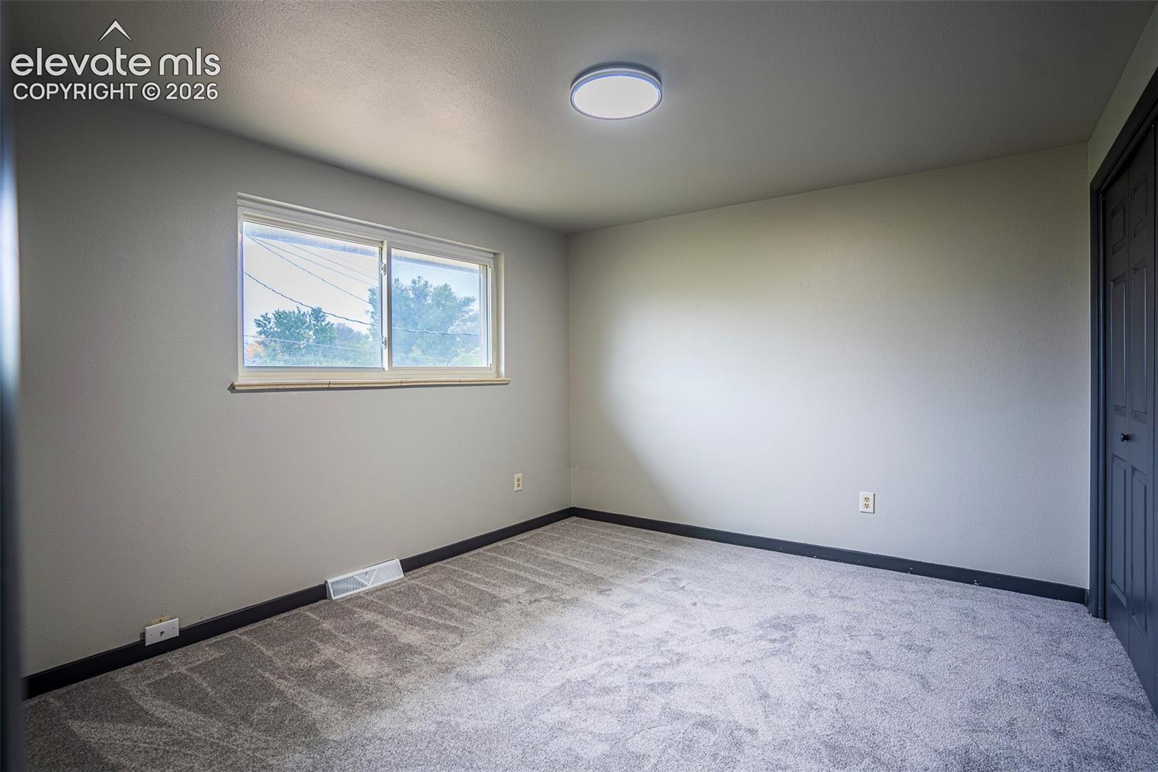 Neutral-toned interior space featuring light grey wall-to-wall carpeting, a sliding-pane window, flush-mount circular lighting, and a dark-finish door