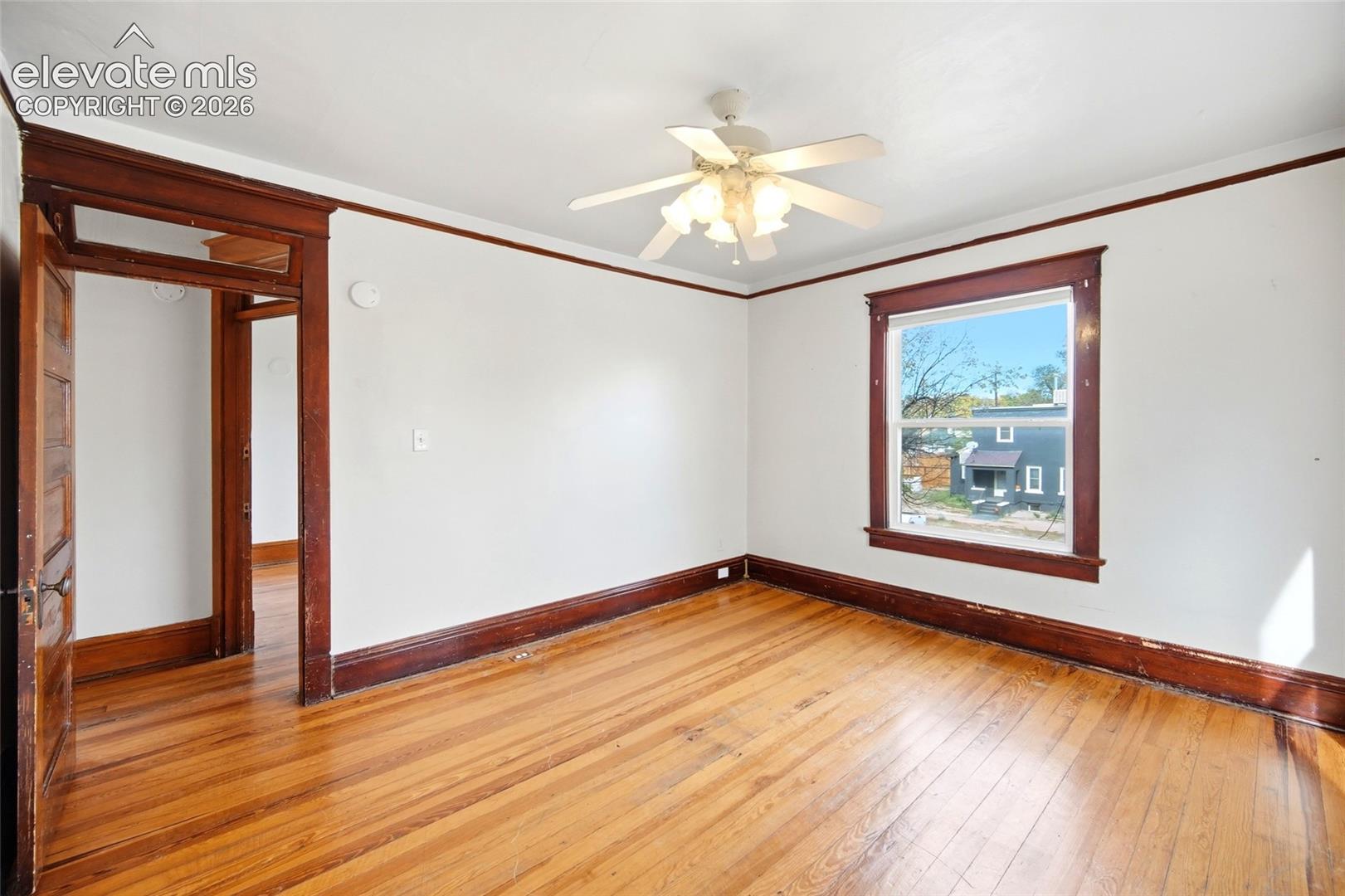 Room featuring wood-finish flooring, dark-stained wood trim, multi-pane window, and ceiling-mounted light fixture with fan