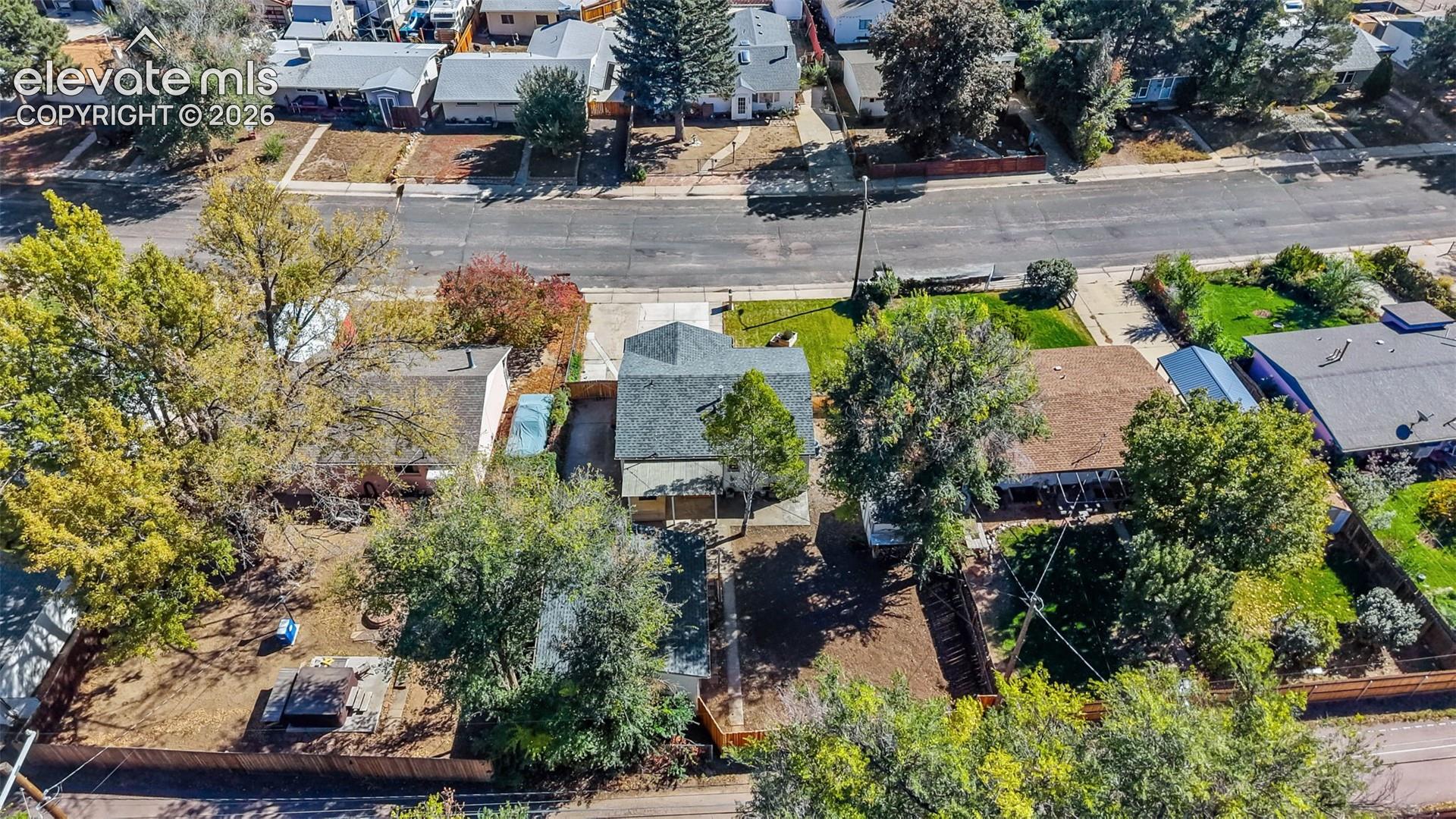 Aerial view featuring a shingled roof, concrete driveway, fenced perimeter, and mature trees on a lot with established landscaping