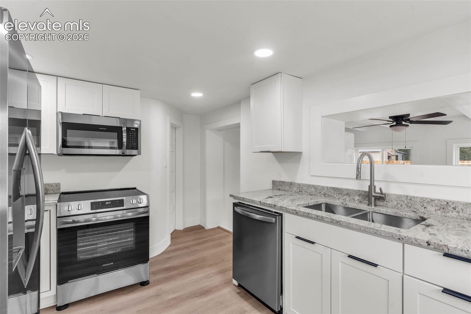 Kitchen featuring white shaker-style cabinetry, stainless steel appliances, stone-surface countertops, dual-basin under-mount sink with pull-down faucet, and wood-finish flooring