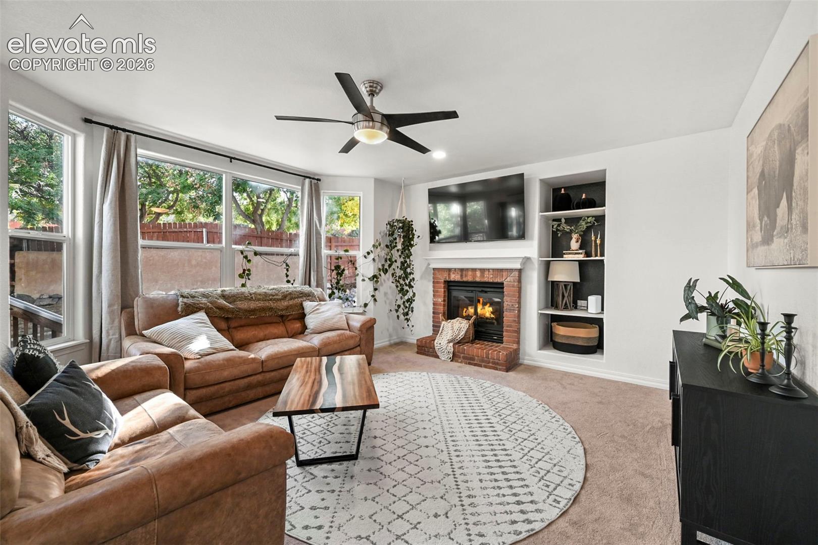 Spacious living area featuring a brick fireplace, built-in shelving, expansive windows, and a ceiling fan