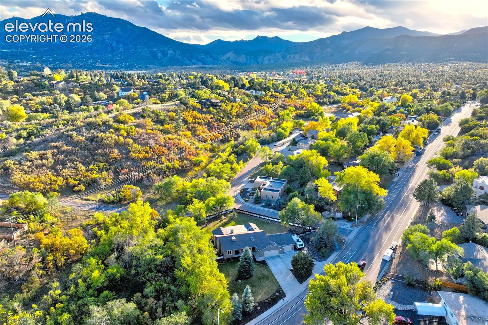 Aerial perspective featuring a multi-level residential structure, concrete driveway, mature evergreen trees, and a mountain range backdrop