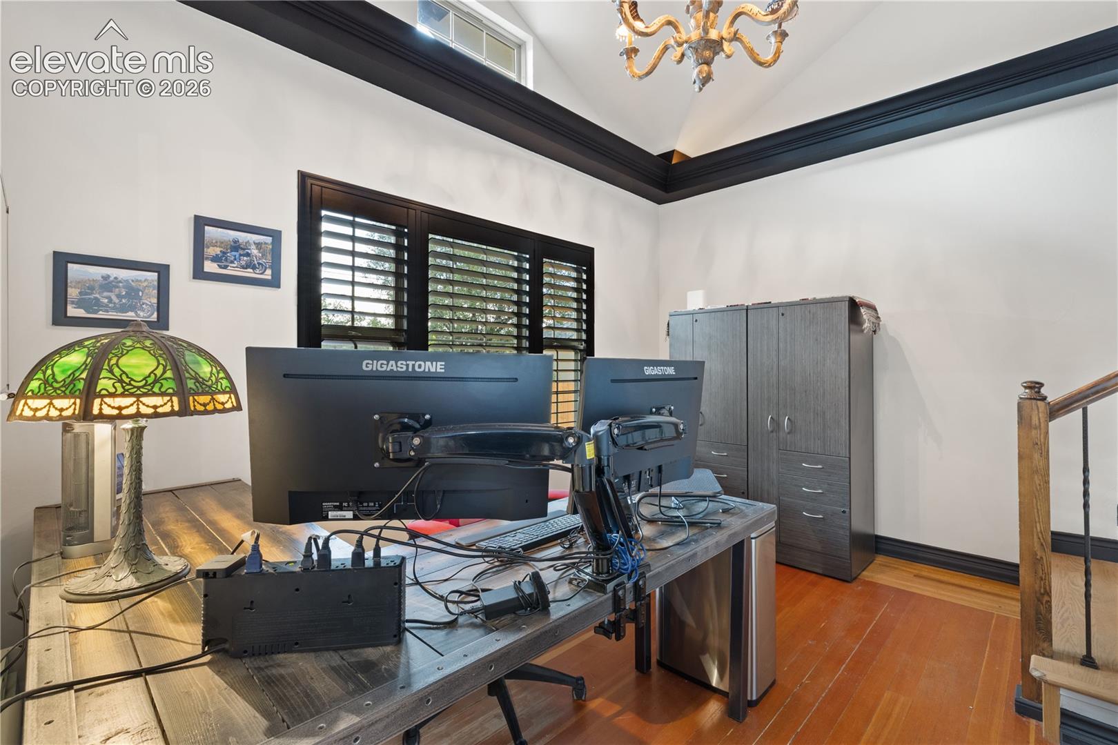 Vaulted ceiling with crown molding, natural wood flooring, window with interior shutters, and a chandelier