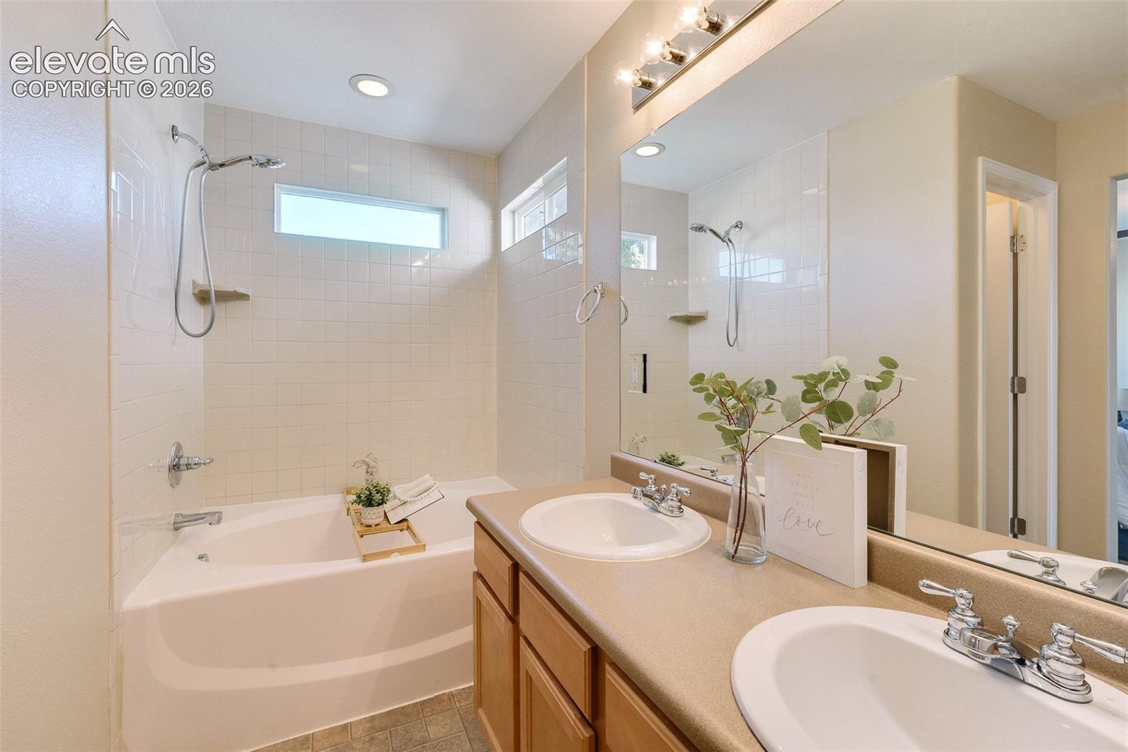 Bathroom featuring a dual-sink vanity with light-tone cabinetry, a full-width mirror, soaking tub with tile surround, and a wall-mounted shower fixture