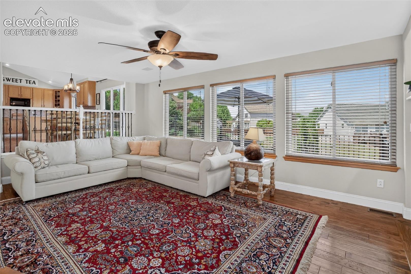 Living area with wood-finish flooring, multiple windows, a ceiling fan, and a partial view of the kitchen with wood cabinetry and built-in appliances