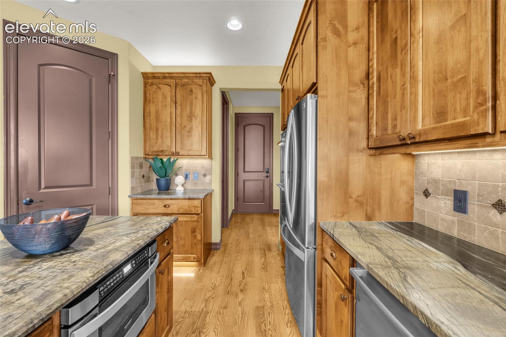 Kitchen featuring wood cabinetry, natural stone countertops, square-tile backsplash, wood-finish flooring, and stainless steel appliances