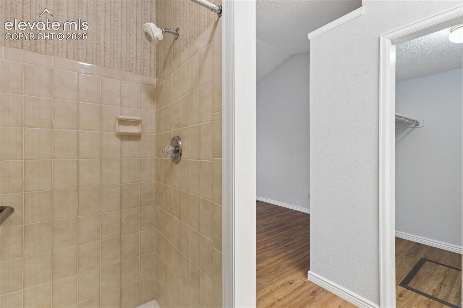 Shower stall featuring square ceramic wall tiling, built-in soap dish, and wall-mounted shower head, finished with wood-finish flooring in the surrounding space and a built-in closet