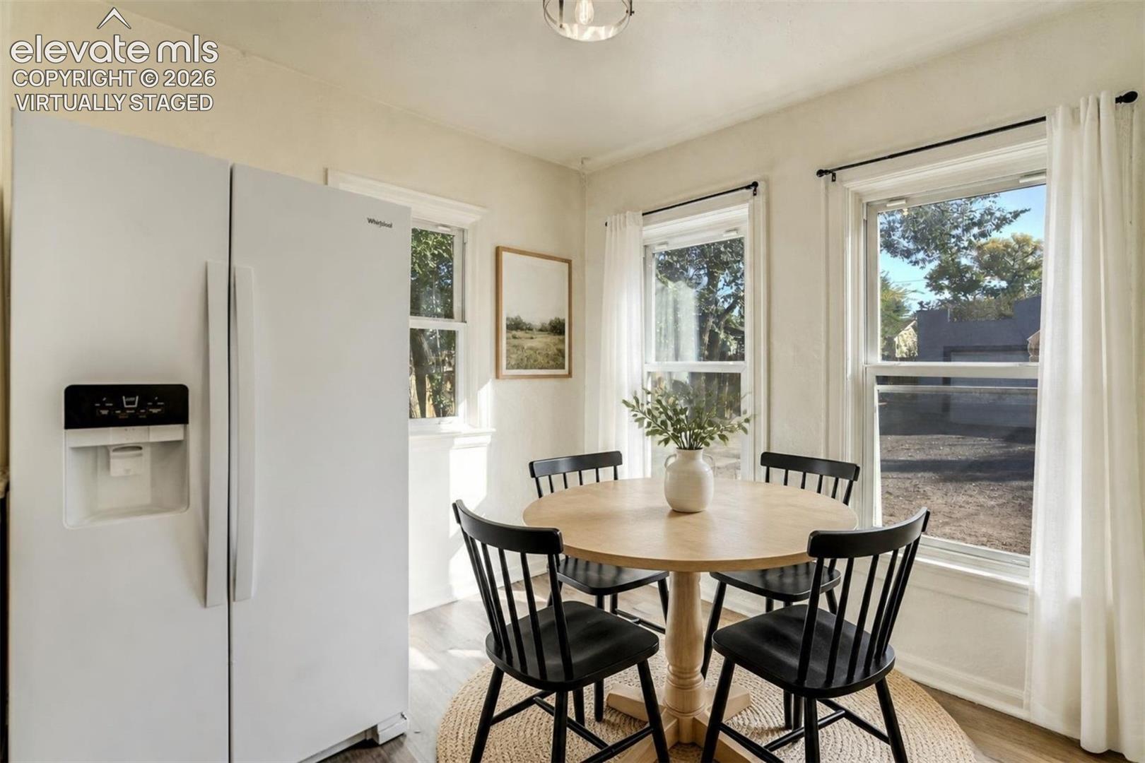 Virtually Staged.  Three windows with white trim, wood-finish flooring, and a built-in white refrigerator with ice dispenser