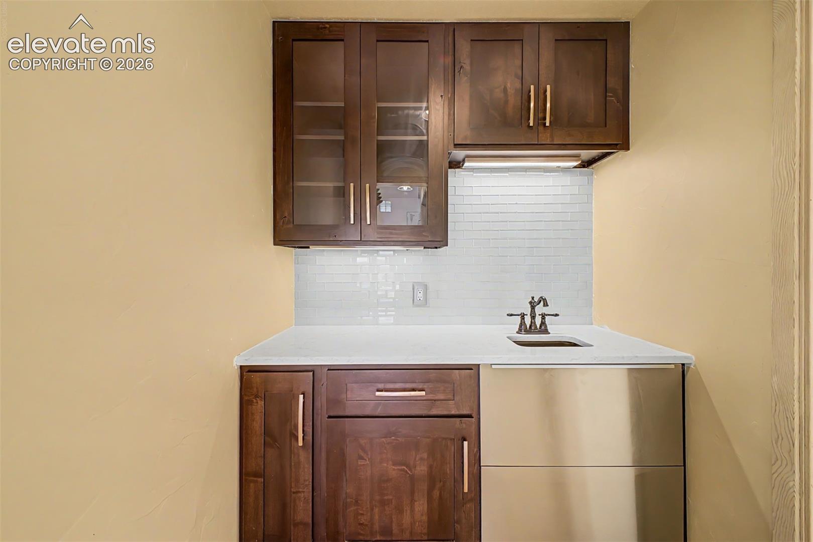 Wet bar featuring wood-finish cabinetry, glass-front upper cabinets, a white countertop with an undermount sink, and a subway tile backsplash