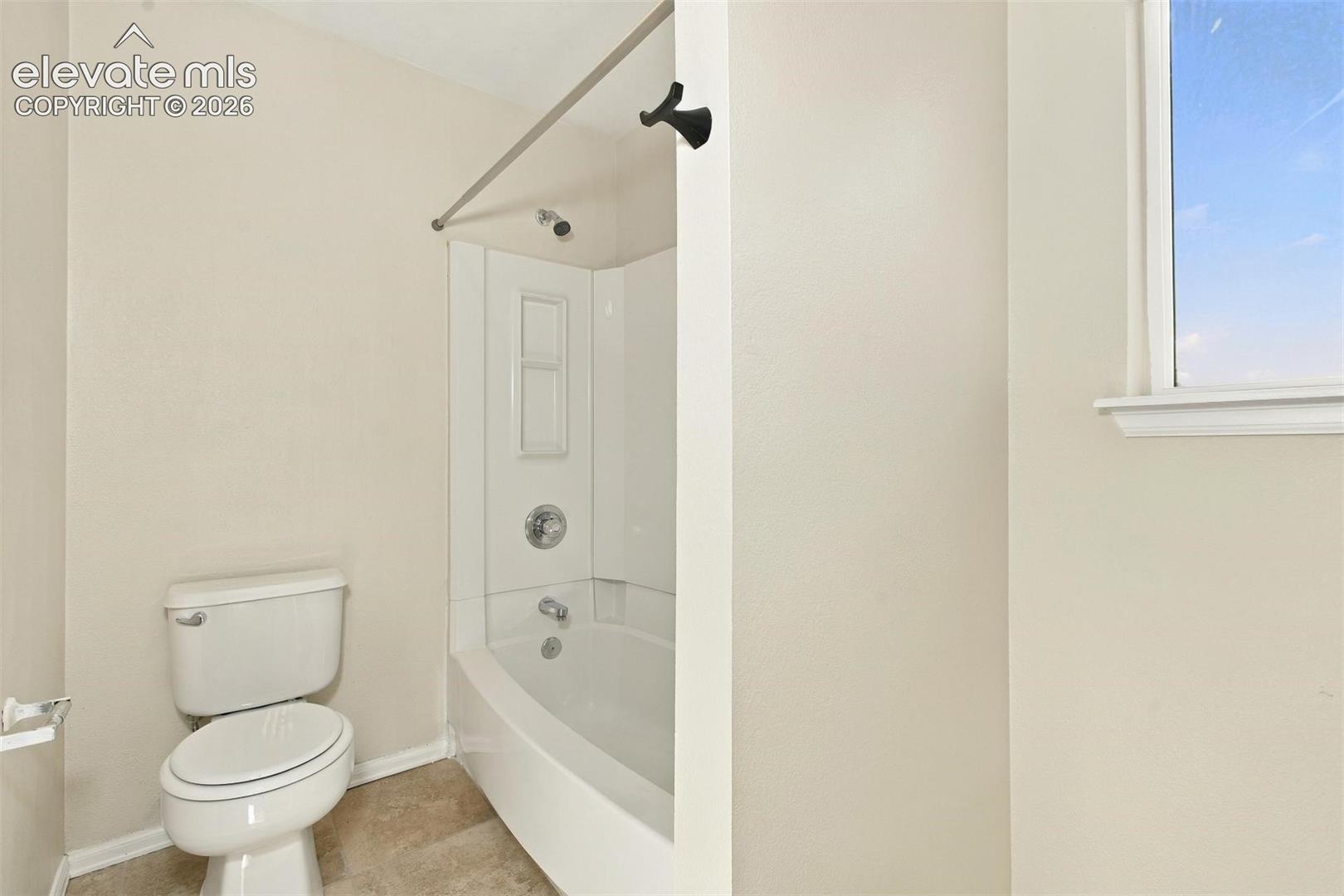 Bathroom featuring a tub-shower combination with integrated shelving, a white toilet, neutral wall paint, and a window with exterior exposure