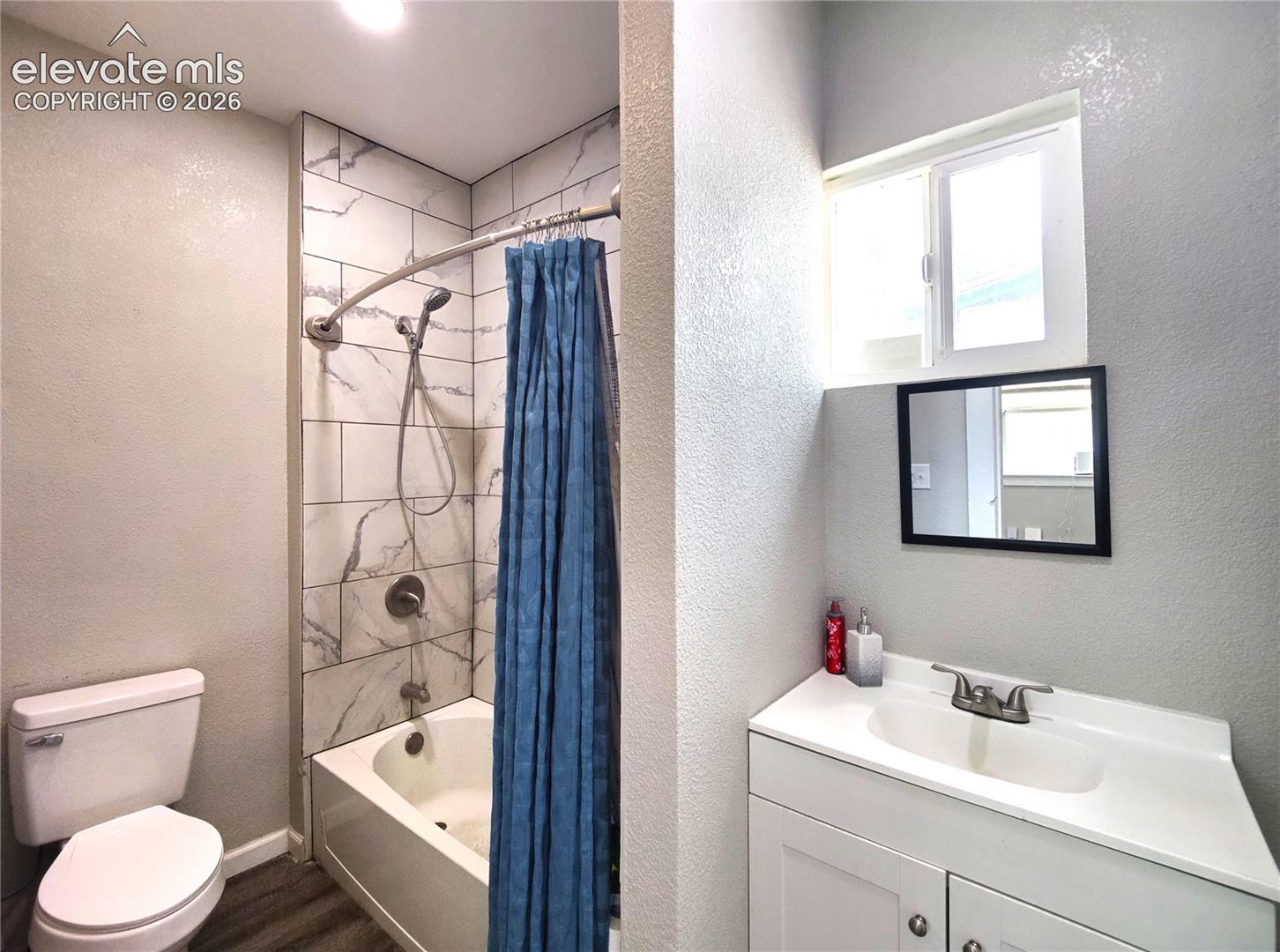 Bathroom featuring a white vanity with integrated sink, a rectangular mirror, a window, a white toilet, and a bathtub with tiled surround