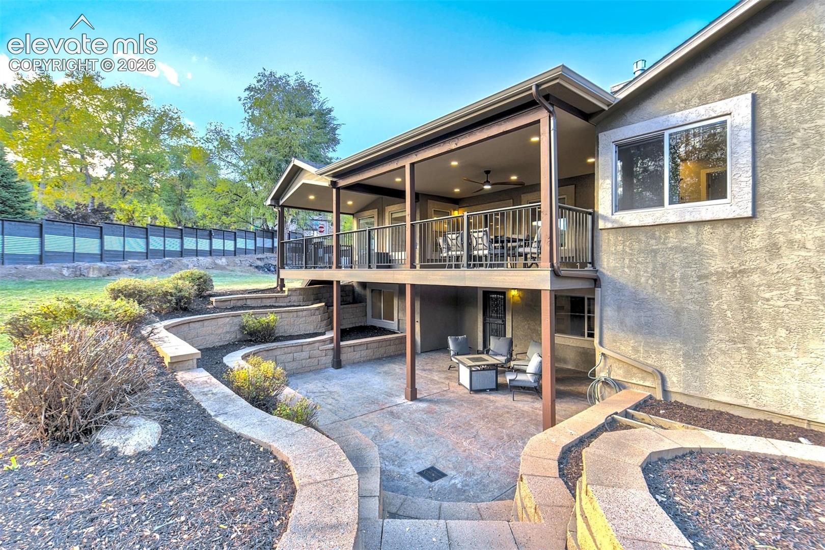Covered rear deck featuring metal railings and recessed lighting, paired with a lower-level concrete patio and tiered stone retaining walls