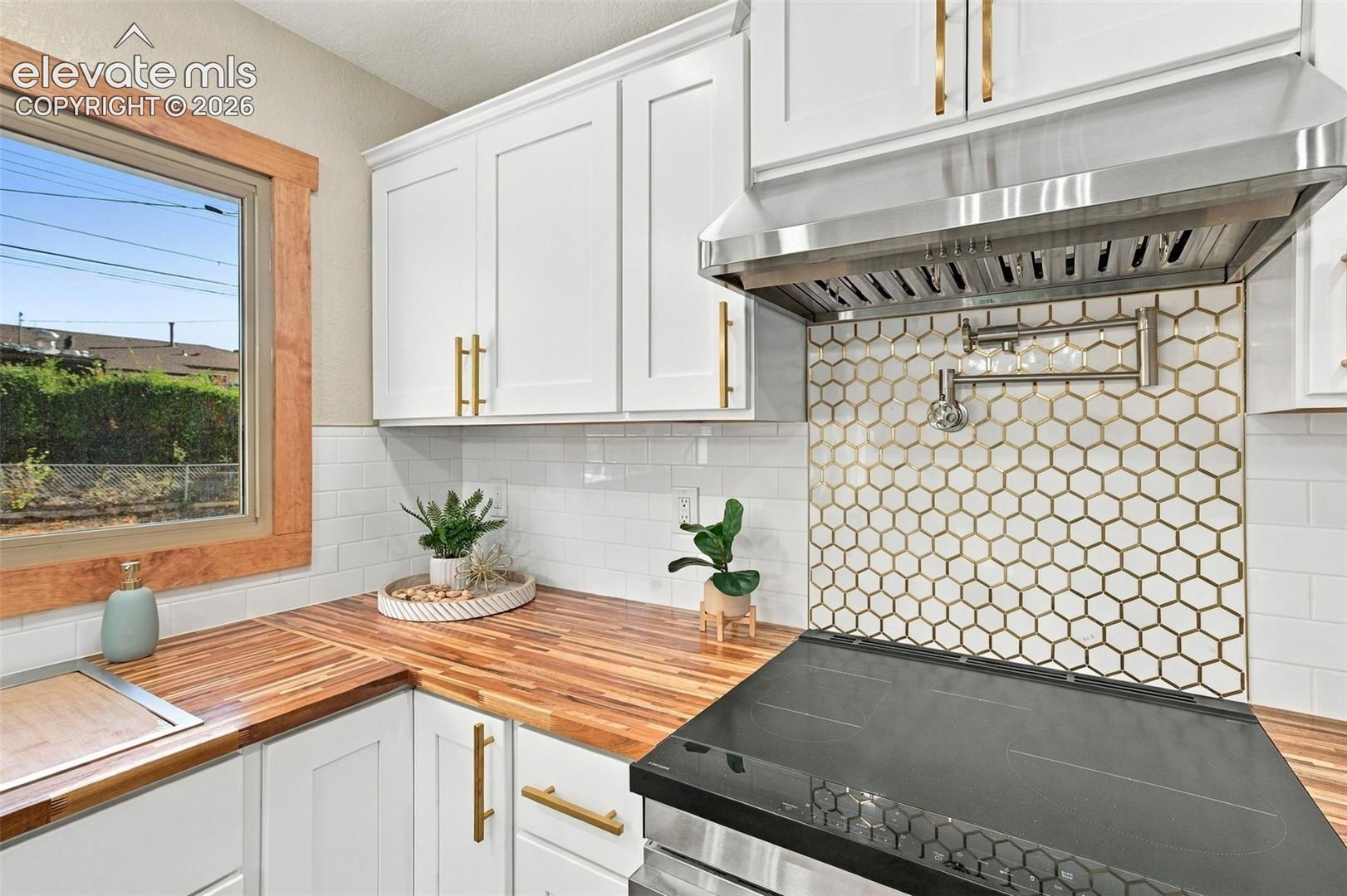 White shaker-style cabinetry with gold-tone pulls, wood-finish countertops, stainless steel range hood, hexagon-pattern tile backsplash, and a wall-mounted pot filler