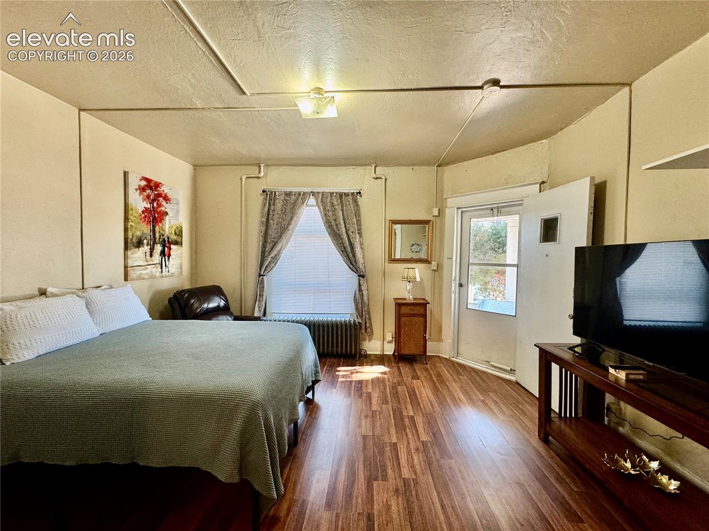 Spacious bedroom featuring wood-finish flooring, a four-poster bed frame, large window with curtains, wall-mounted flat-screen television, and built-in cabinetry