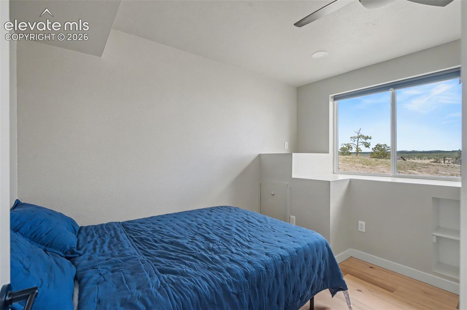 Room interior featuring neutral wall paint, wood-finish flooring, a ceiling fan, a built-in recessed niche, and a window with exterior landscape
