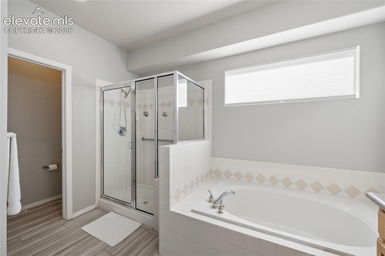 Bathroom featuring a glass-enclosed shower, a tiled soaking tub with a deck-mounted faucet, wood-finish flooring, and a horizontal transom window