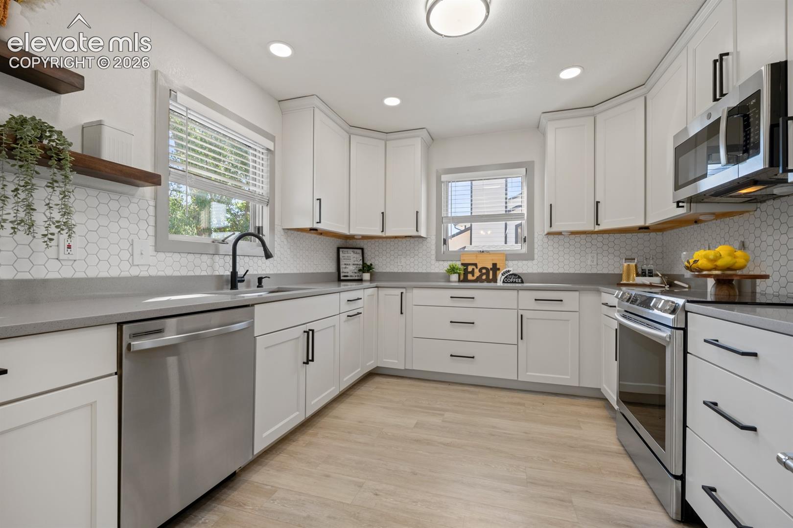 White shaker-style cabinetry with matte black hardware, stainless steel appliances, grey countertops, white hexagonal tile backsplash, and light wood-finish flooring