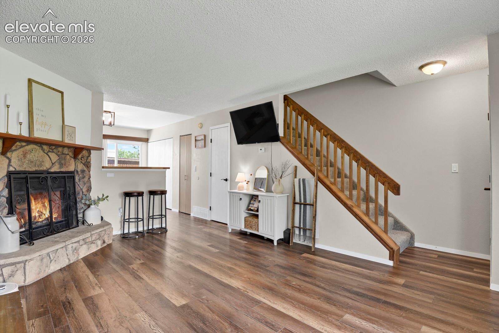 Stone-clad fireplace with a wood mantle, wood-finish flooring, a built-in breakfast bar, and a carpeted staircase with wood banister