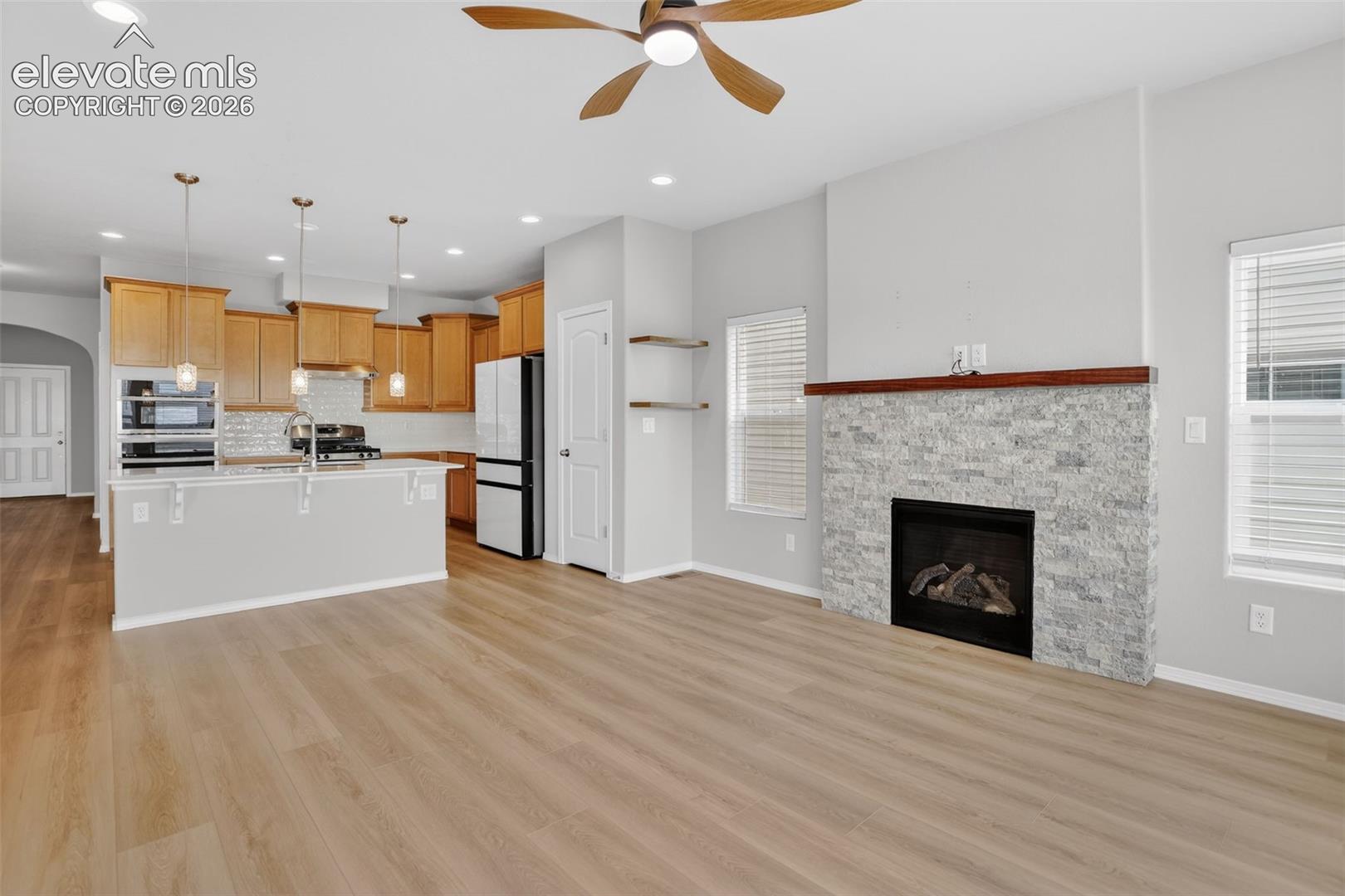 Open-concept living space featuring wood-finish flooring, a contemporary fireplace with stacked stone surround, and a kitchen island with pendant lighting