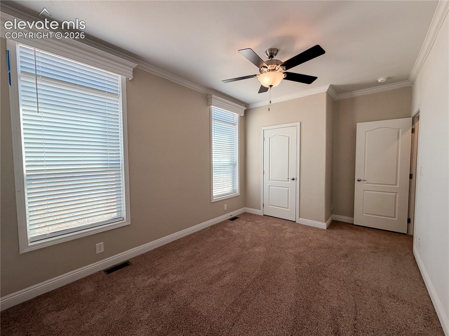 Neutral-toned interior featuring plush carpeting, crown molding, multi-panel doors, dual windows with fitted blinds, and a ceiling-mounted fan with light fixture