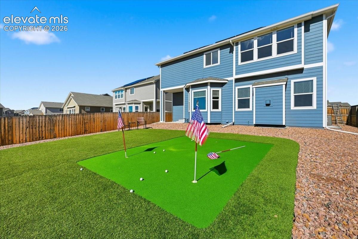 Rear exterior featuring blue horizontal siding, white trim, a built-in putting green, synthetic turf lawn, and perimeter wood privacy fencing