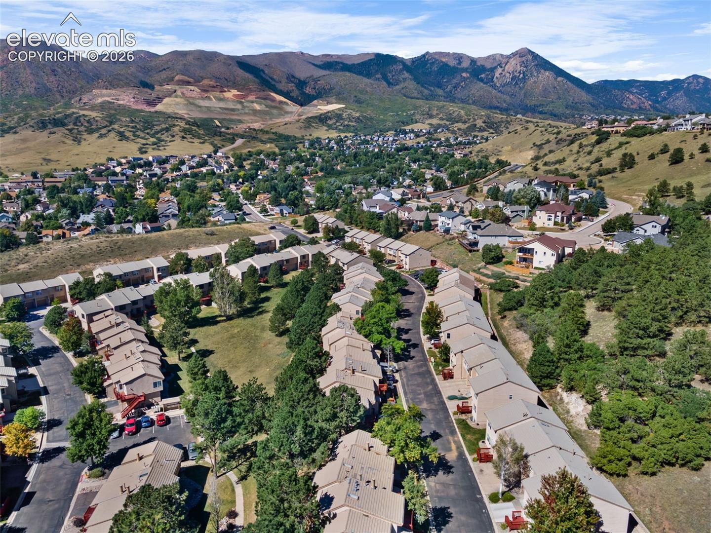 Aerial perspective of a multi-unit residential community featuring tiered shingle roofing, paved roadways, mature evergreen landscaping, and a prominent mountain range backdrop
