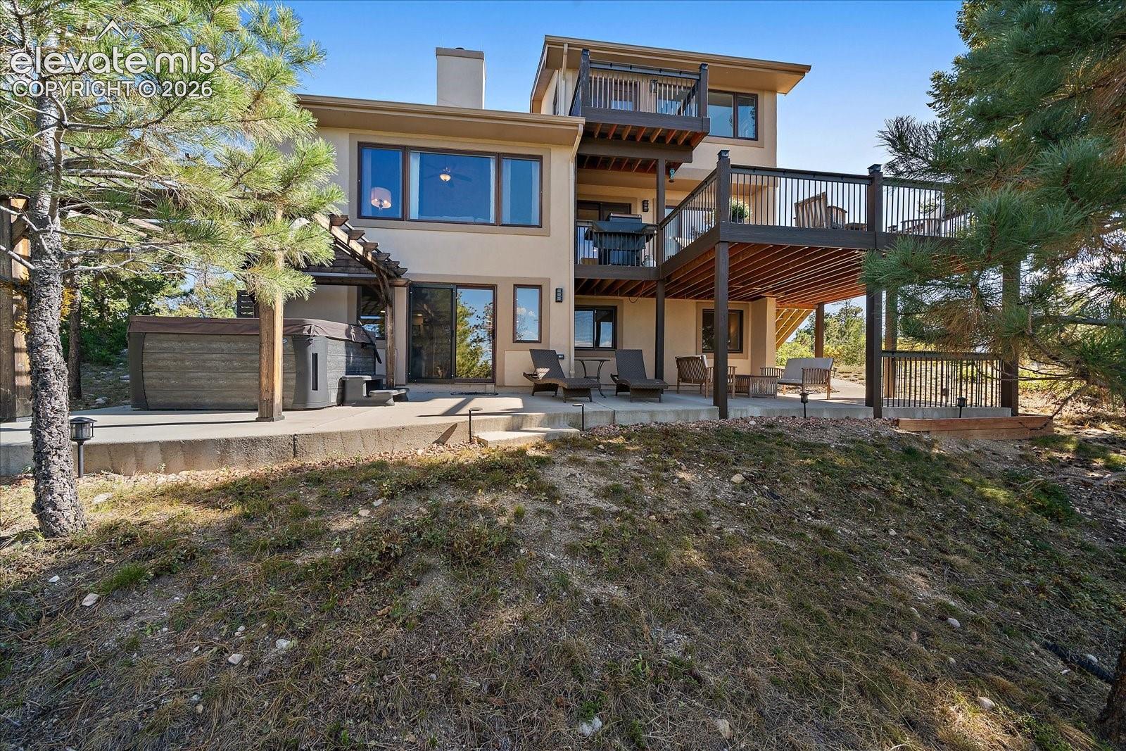 Multi-level home exterior featuring a raised wood deck with metal railings, a concrete ground-level patio, built-in hot tub, and a wooden pergola