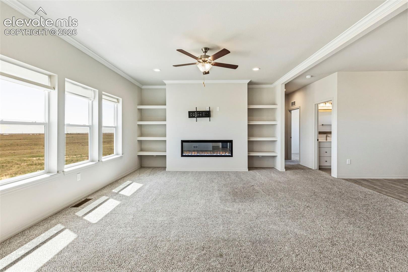 Spacious living area featuring a built-in electric fireplace with mounted shelving, three large windows, a ceiling fan, and crown molding