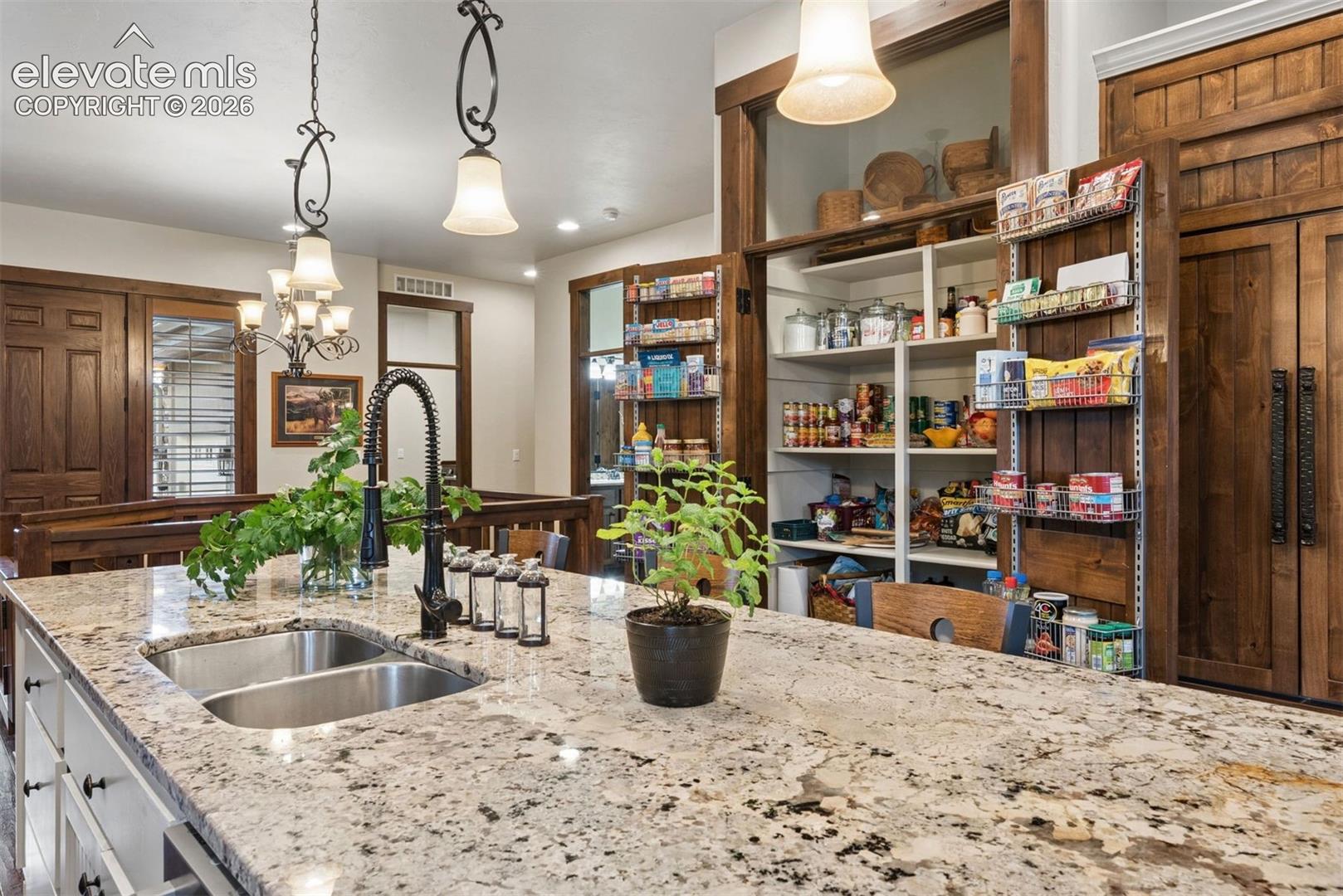 Kitchen island featuring stone countertops, an undermount double-basin sink, a matte black high-arch faucet, custom wood cabinetry, and a walk-in pantry with built-in shelving