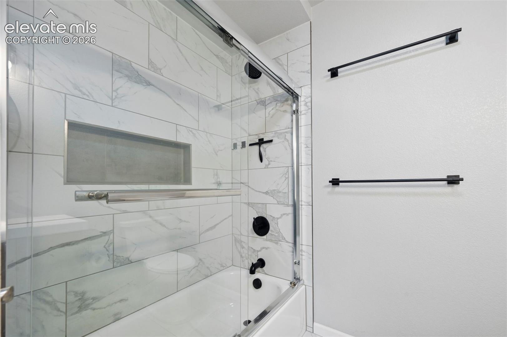 Full bathroom on main adjacent to the secondary bedroom on main.  Bathtub featuring large-format stone-look tile surround, built-in recessed wall niche, sliding glass enclosure, and matte black fixtures